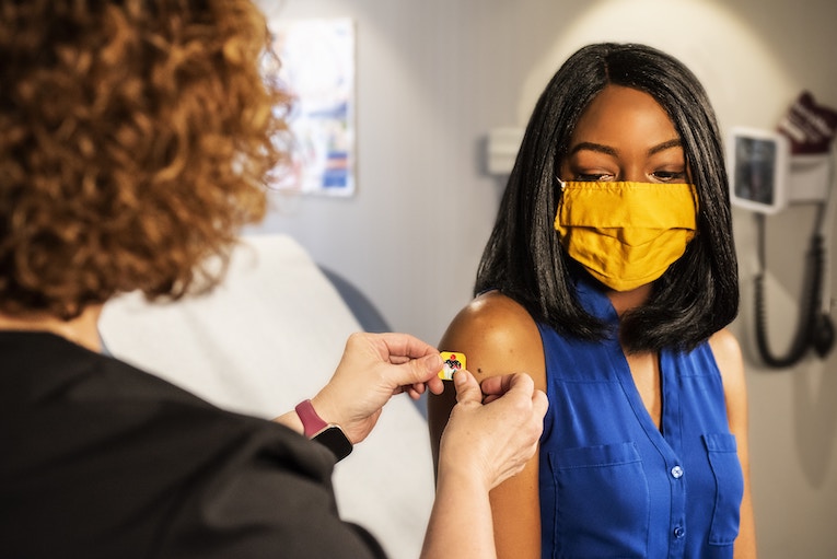 back of person putting band-aid on the arm of another person wearing medical mask