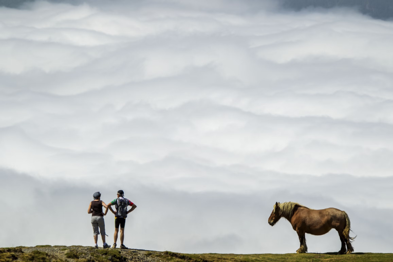 two people and horse standing on mountain looking down at clouds