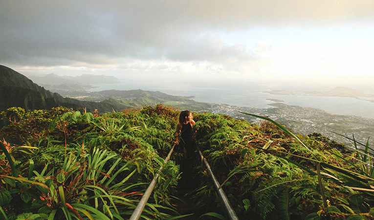 woman hiking up hillside covered in green tropical plants under a light but cloudy sky