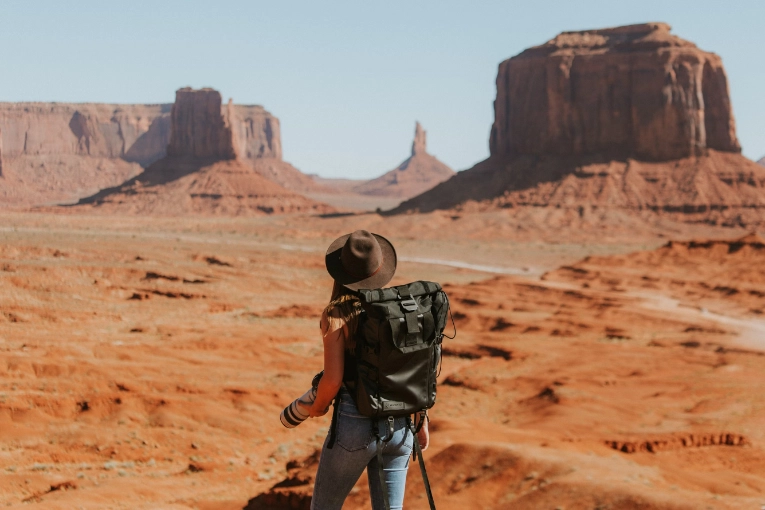 person wearing backpack and holding camera while standing on brown sand looking at brown rock formations