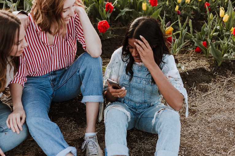 three people sitting in front of flowering and laughing while looking at smartphone one is holding