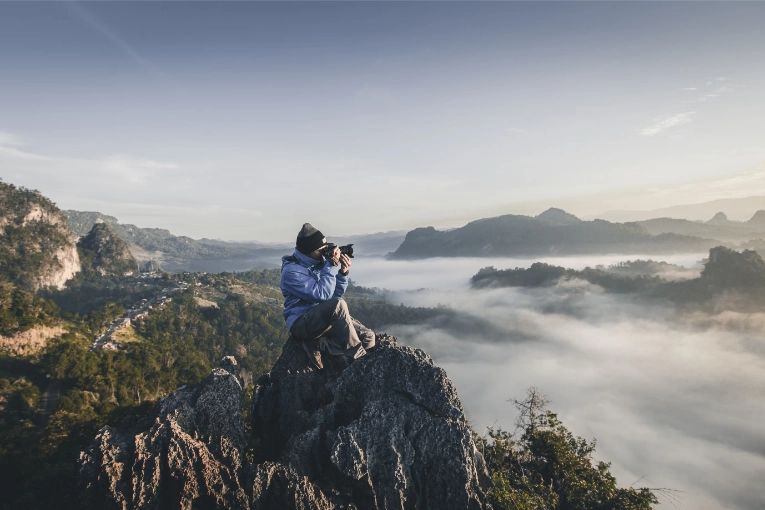 person holding camera while sitting on mountain top above clouds