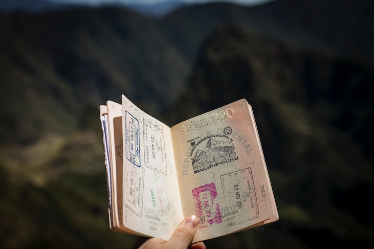 person holding passport open to page with stamps with mountains blurred in background