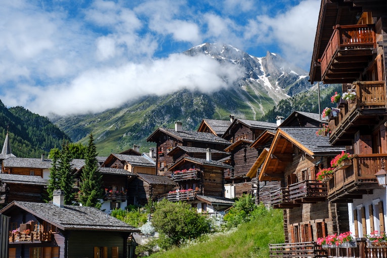 A village of wooden log cabins sits at the base of a snow peaked mountain range in Switzerland