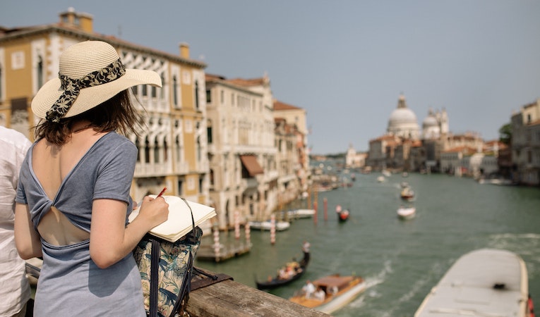 expat in blue shirt and brown hat standing on a brown wooden bridge overlooking a canal in venice