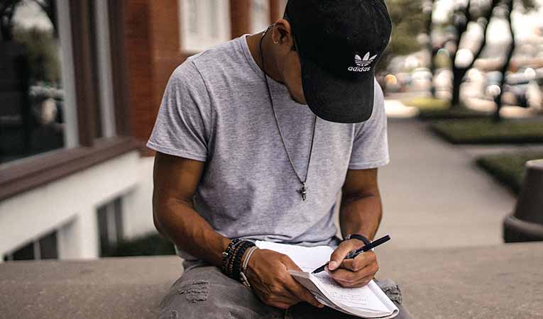 Teen boy in black adidas cap writing in notebook/working on homework