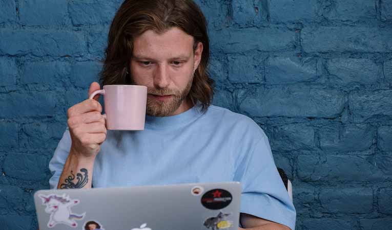 person using a laptop against a blue brick wall while drinking a cup of coffee 