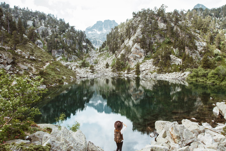 person standing near water surrounded by mountains with rocks and trees