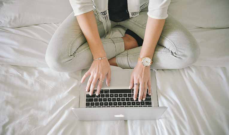 shot from above, woman in white sitting on white bedspread with laptop typing