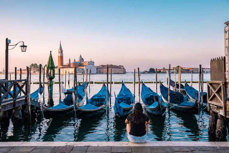 person sitting in front of water and boats with buildings across the water in the distance