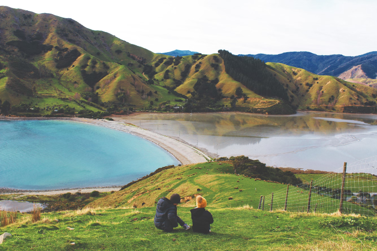 two people sitting on grassy hill overlooking body of water and mountains