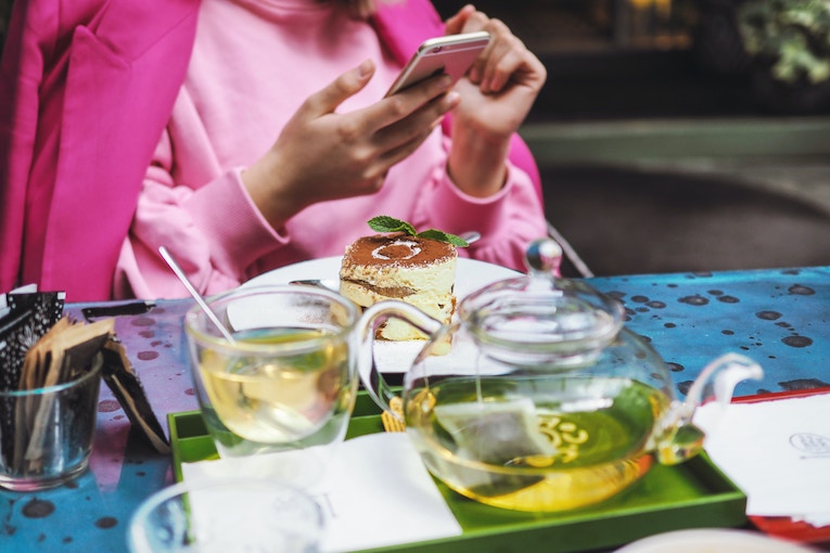 person behind a table holding iPhone with tea and pastry on a table