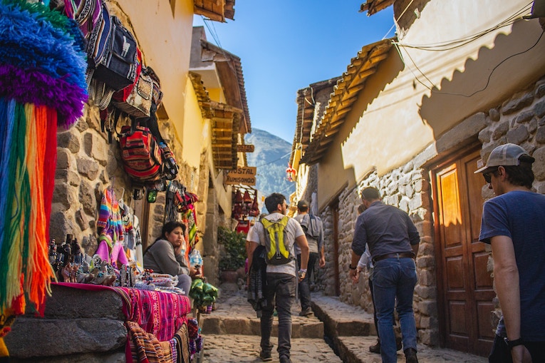 person walking on narrow street with colorful bags hanging and mountain behind